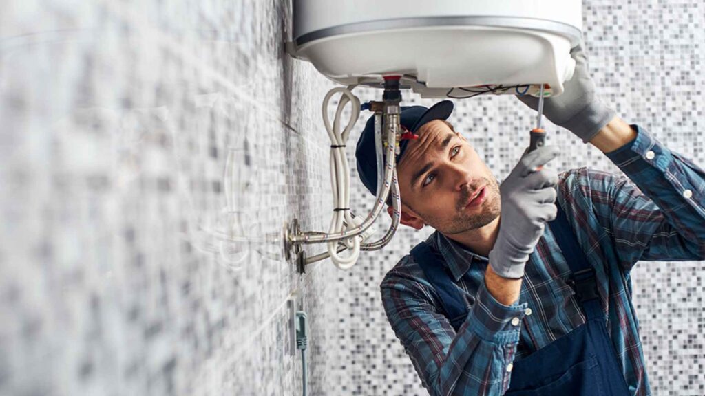 A plumber inspects a heating system in a tiled bathroom, using a flashlight and wrench, wearing gloves and a plaid shirt.