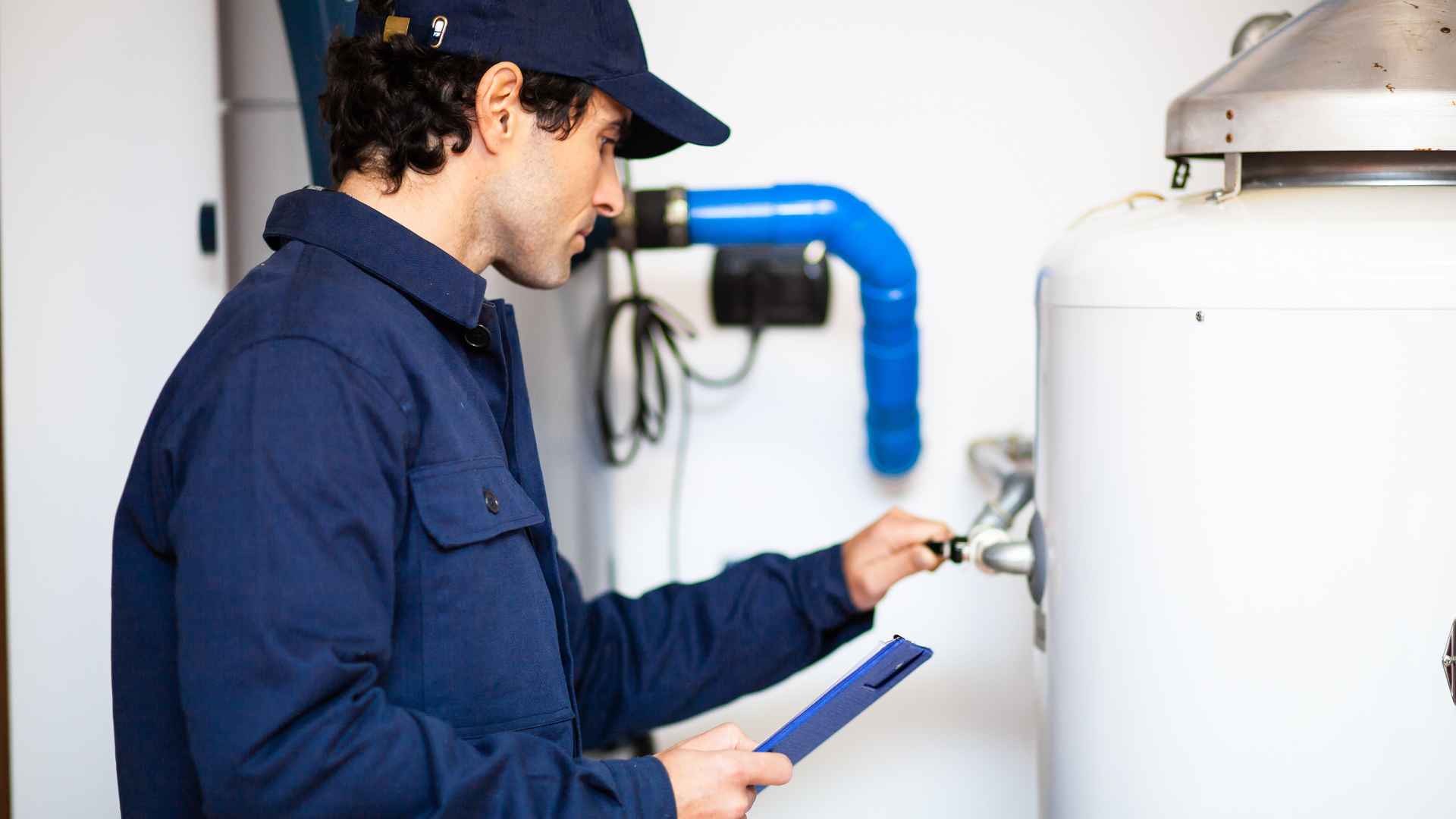 A technician in a navy blue uniform inspecting a large water heater replacement with a clipboard in hand.
