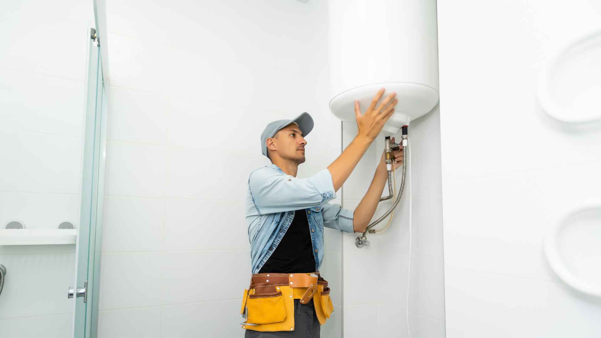 A plumber in a cap and tool belt installs a water heater in a white bathroom.