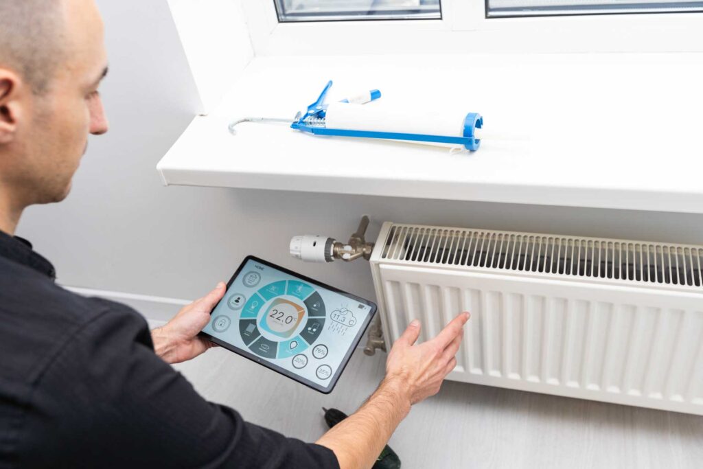 A man adjusts a radiator thermostat using a digital tablet with a heating control app, tools and water softening equipment visible beside him.