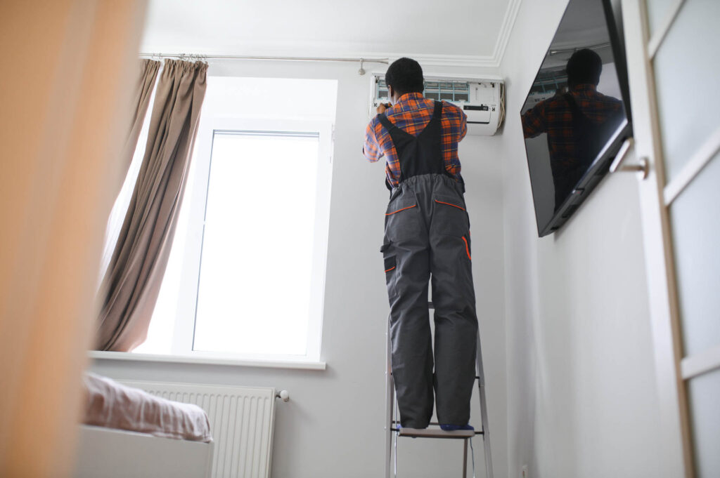 A man on a stepladder installing an air conditioning unit above a door in a bright, modern room after hydro jetting the systems.