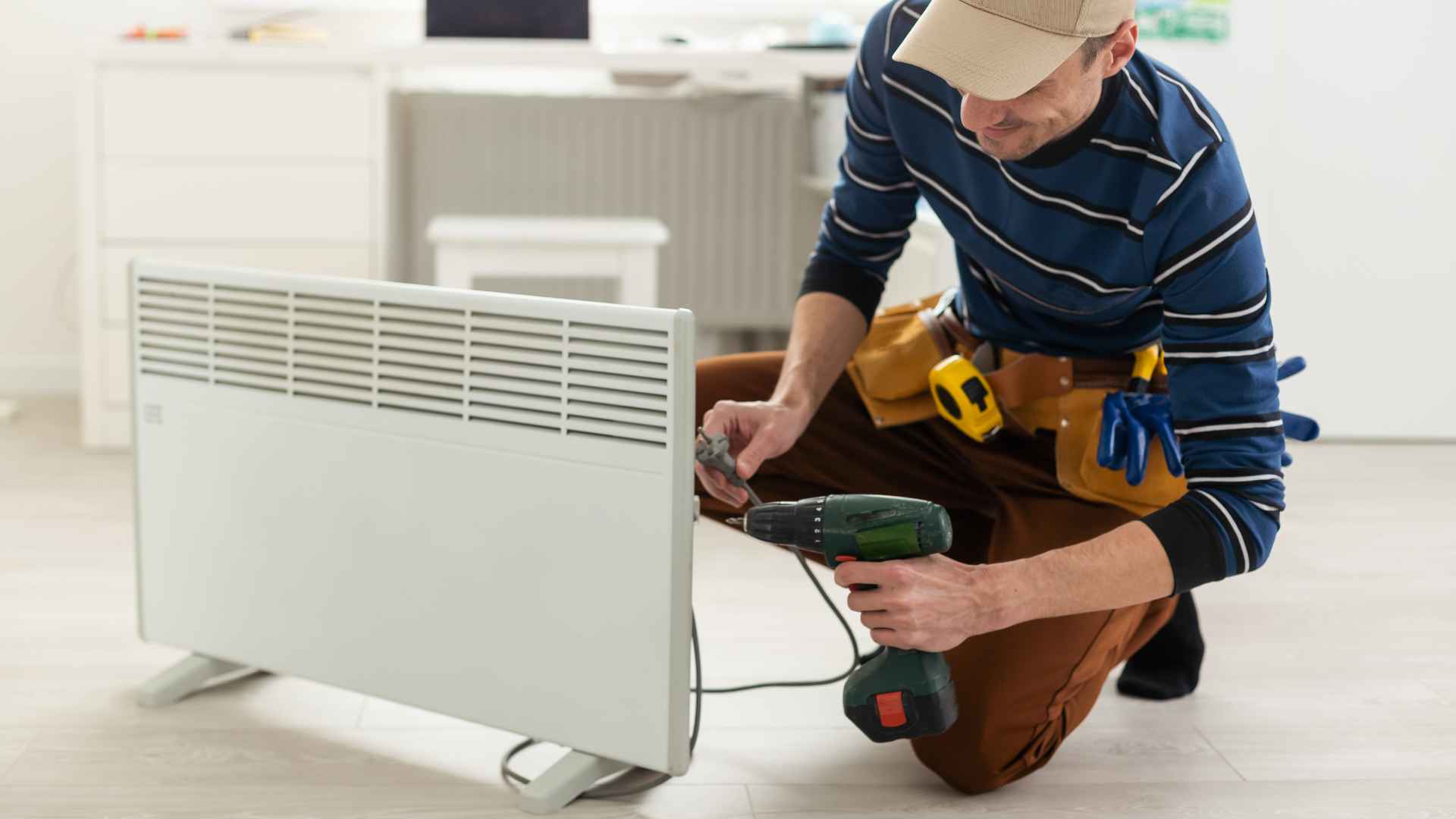 A technician in a striped shirt and cap repairs a white radiator with a cordless drill, attending to the gas line in a bright room.