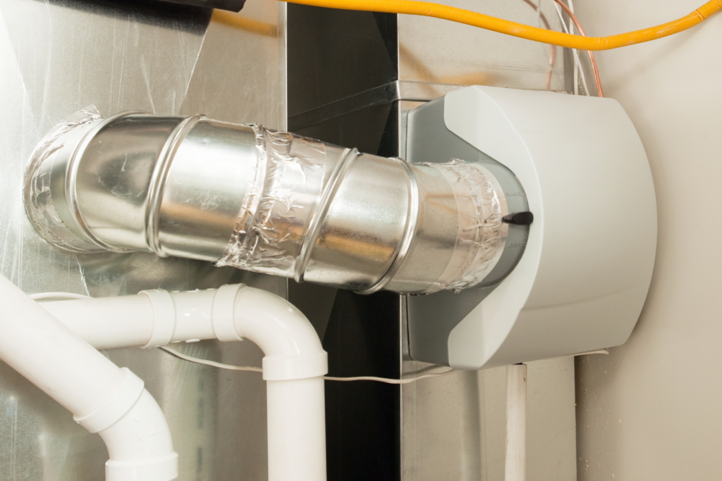 Close-up view of a ductwork connection sealed with foil tape and connected to a grey, wall-mounted HVAC unit, with white water filtration pipes visible.