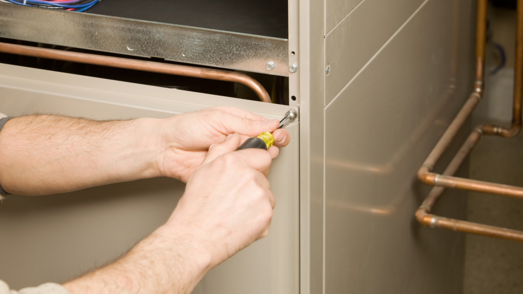 A technician's hands using a screwdriver to connect wiring in an HVAC unit, with copper pipes and water filtration components visible in the background.