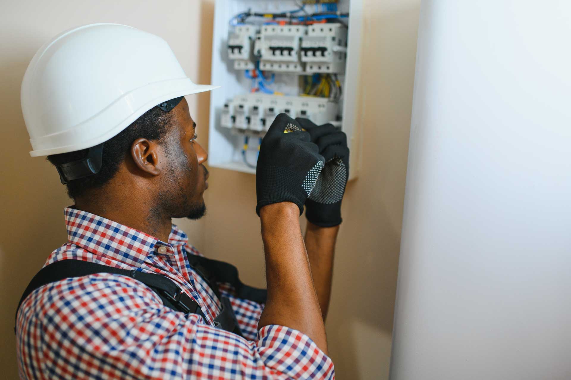 An electrician wearing a hard hat and gloves works on a tub replacement circuit breaker panel.
