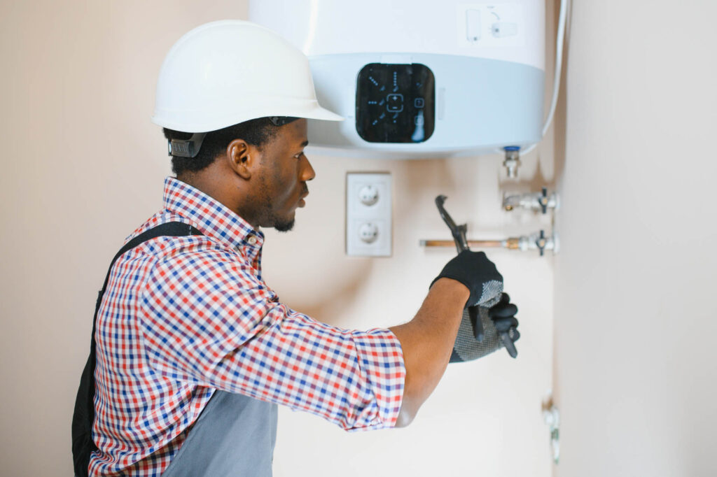 A man in a hard hat and gloves adjusts settings on a wall-mounted water heater using a wrench, carefully checking the gas line.