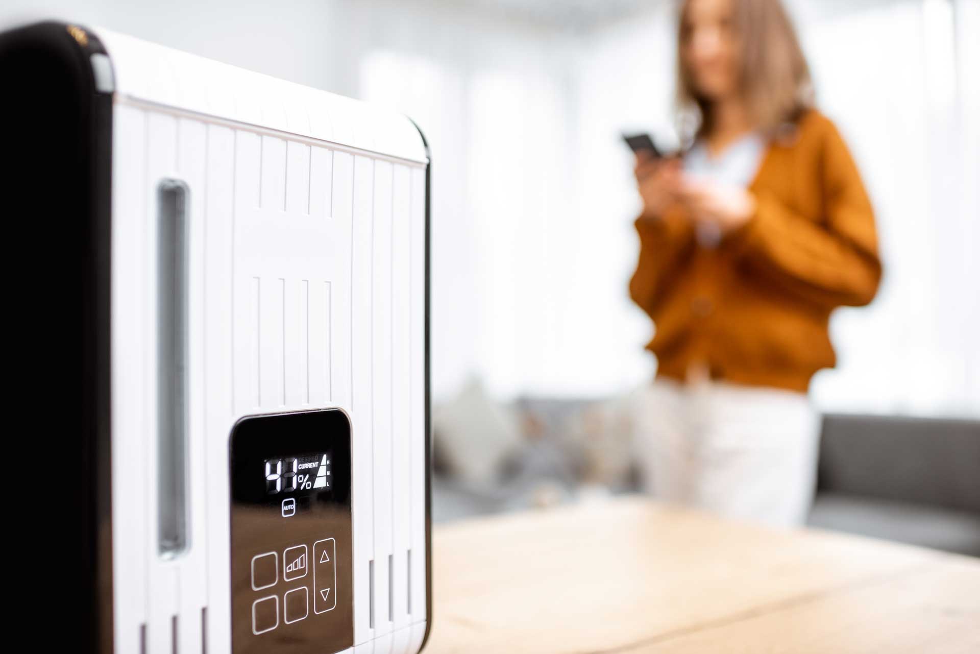 A modern white toaster with a digital display on a kitchen counter, with a blurred woman holding a phone near a water filtration system in the background.