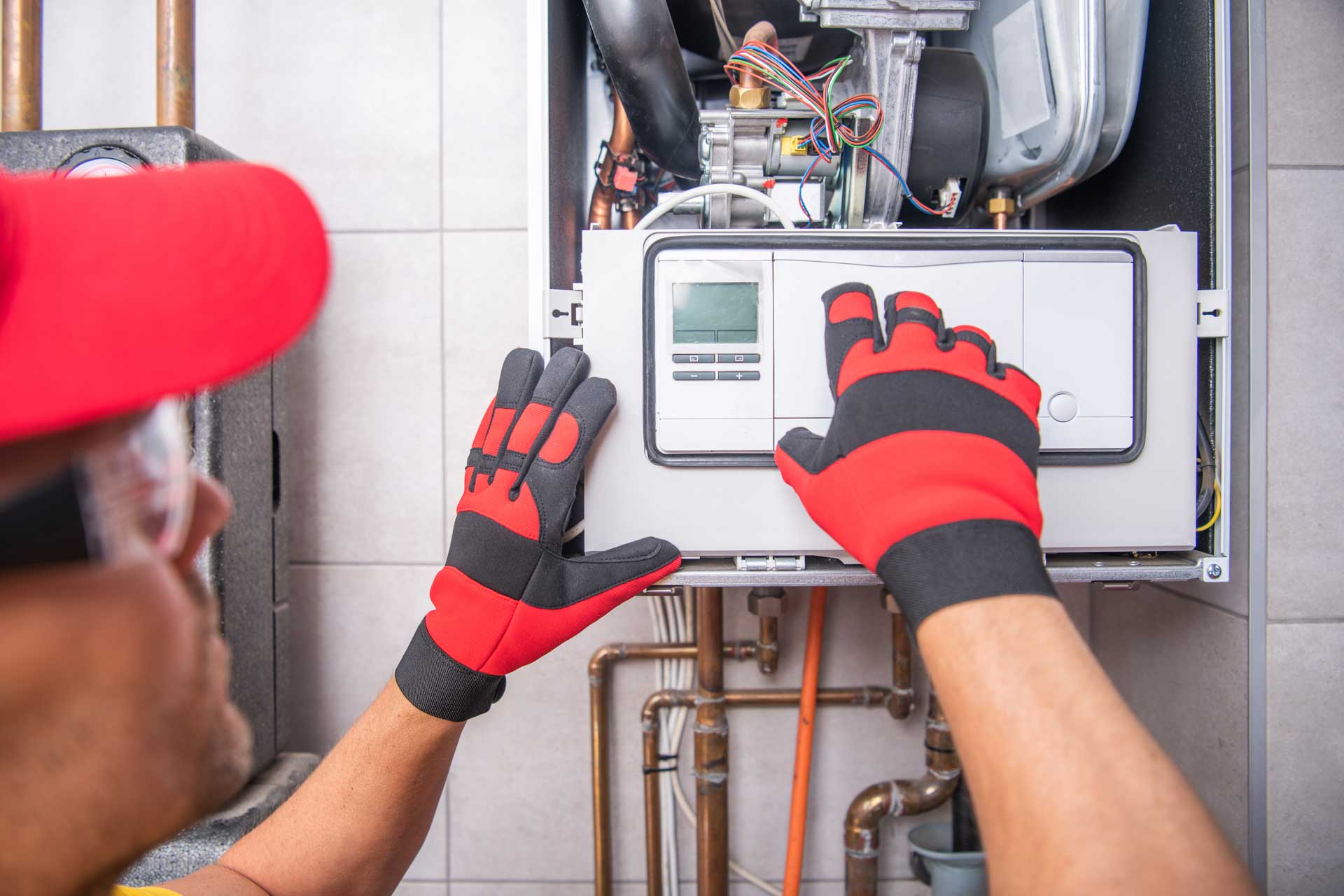 Technician in a red cap and gloves adjusts settings on a residential gas furnace's gas line control panel.