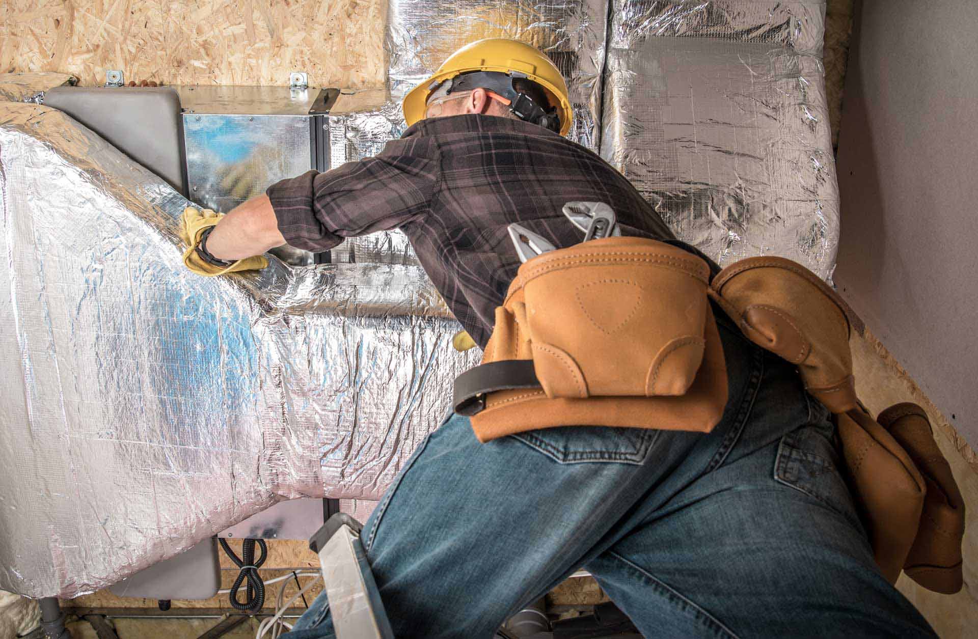 A worker in a hard hat and gloves installs water softening insulation in a wall, wearing a tool belt.
