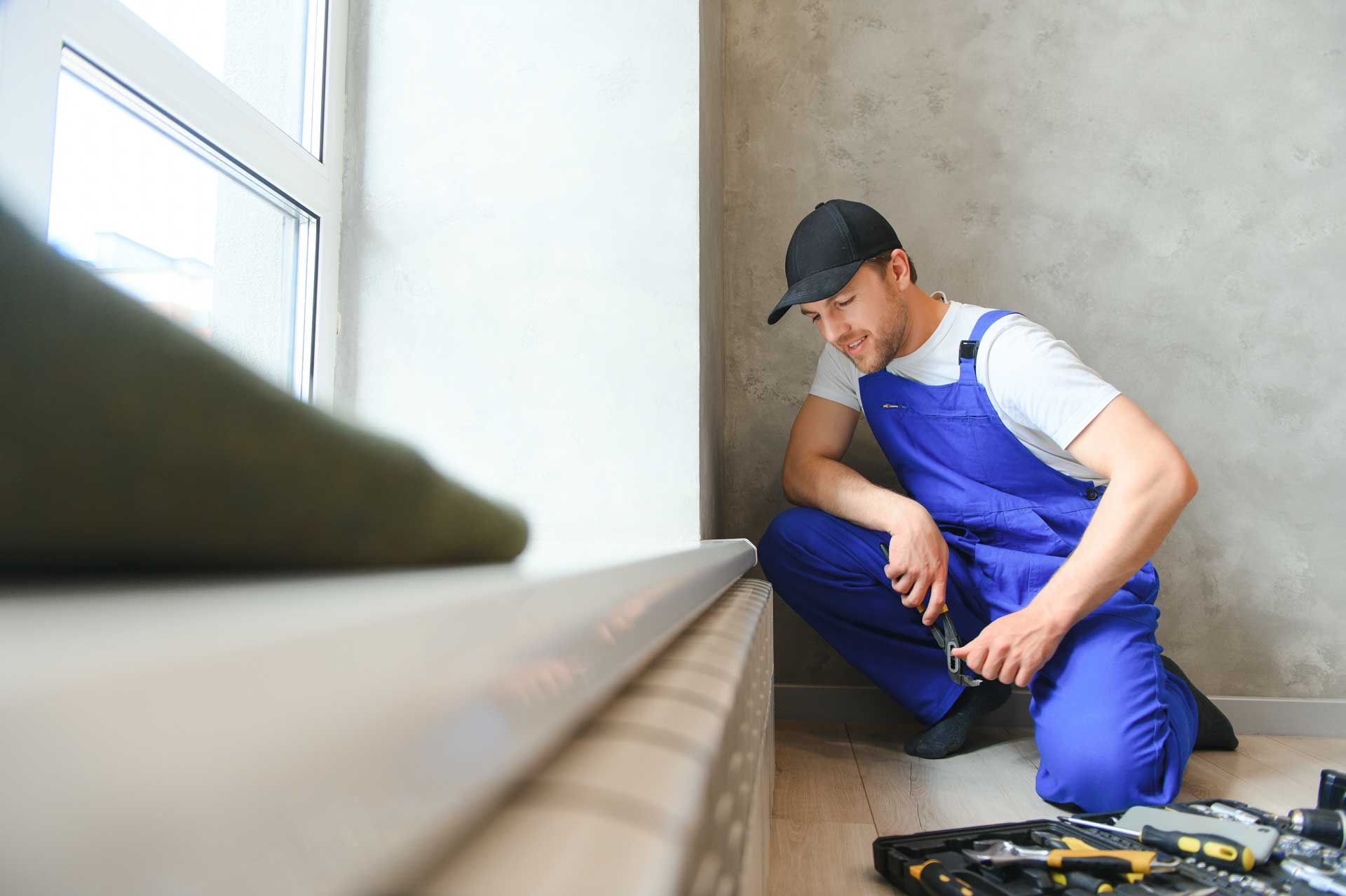 A worker in a blue uniform and cap installs or repairs a gas line in a room with gray walls and a large window.