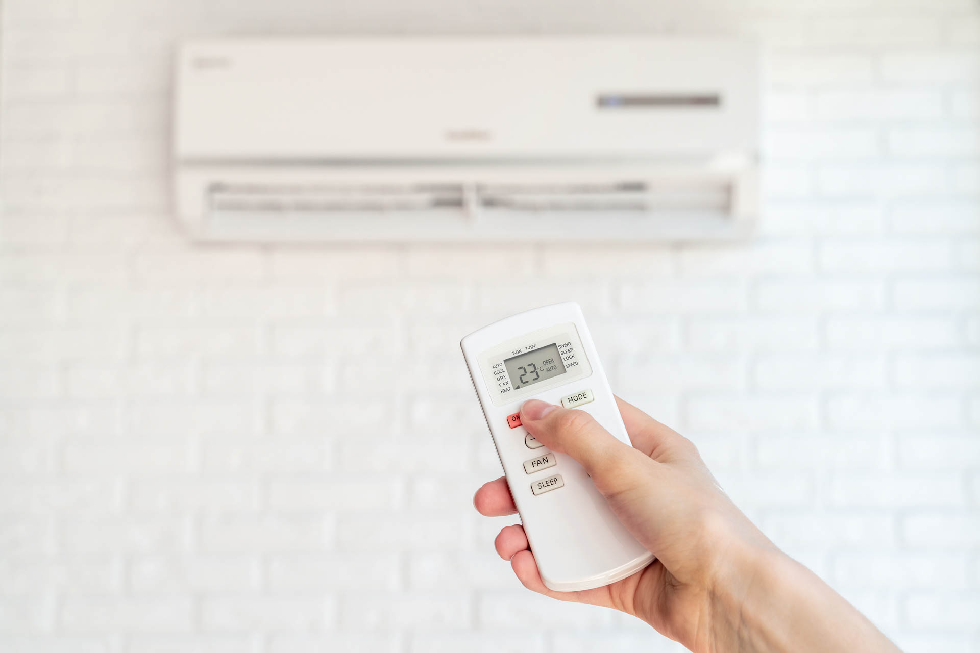 A hand holding a remote control aimed at an air conditioner mounted on a white brick wall, adjusting the temperature to 23 degrees Celsius.