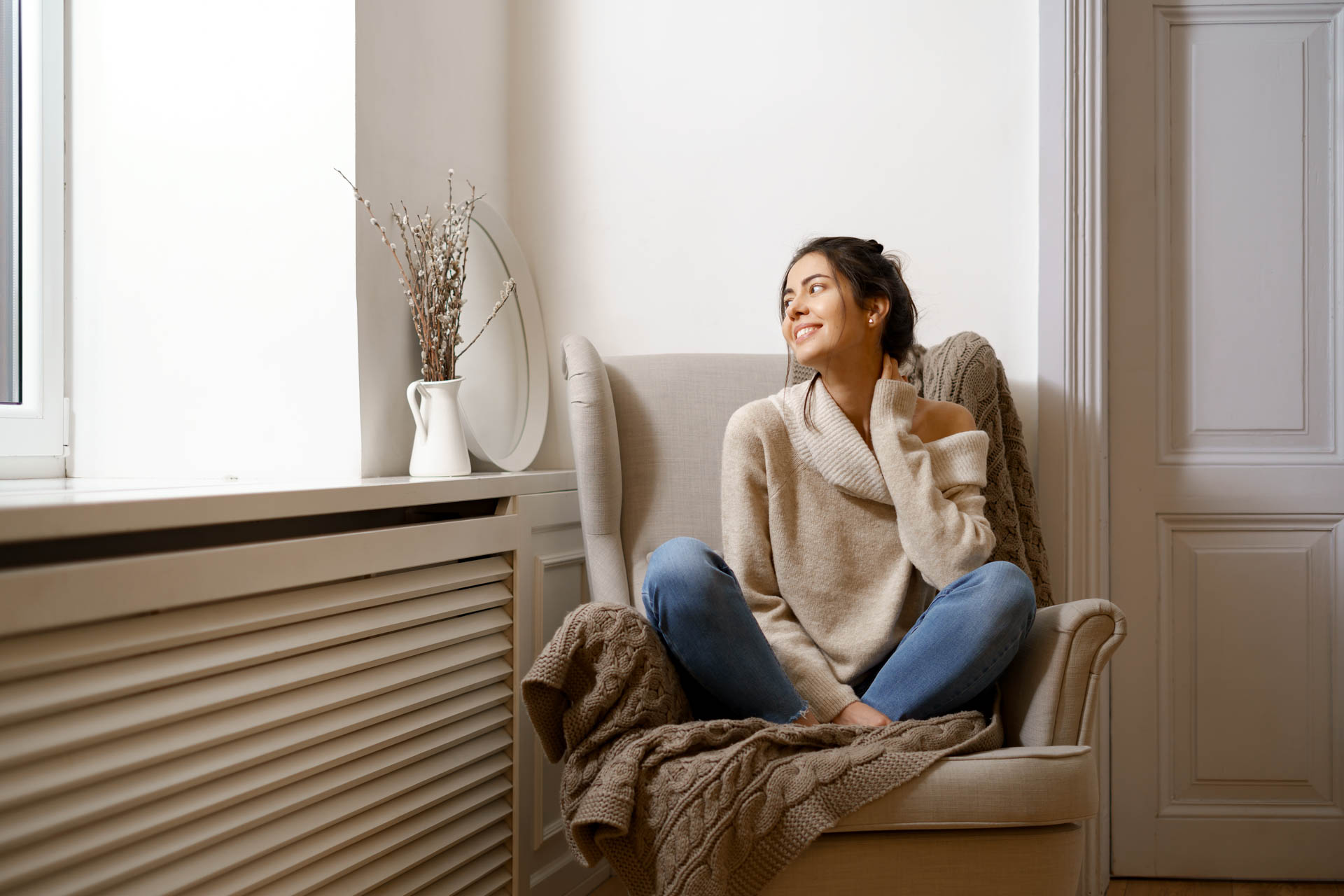 A woman in a cozy sweater sits relaxed in an armchair by a window, smiling and looking away, with a vase of branches and a book on heating and cooling nearby.