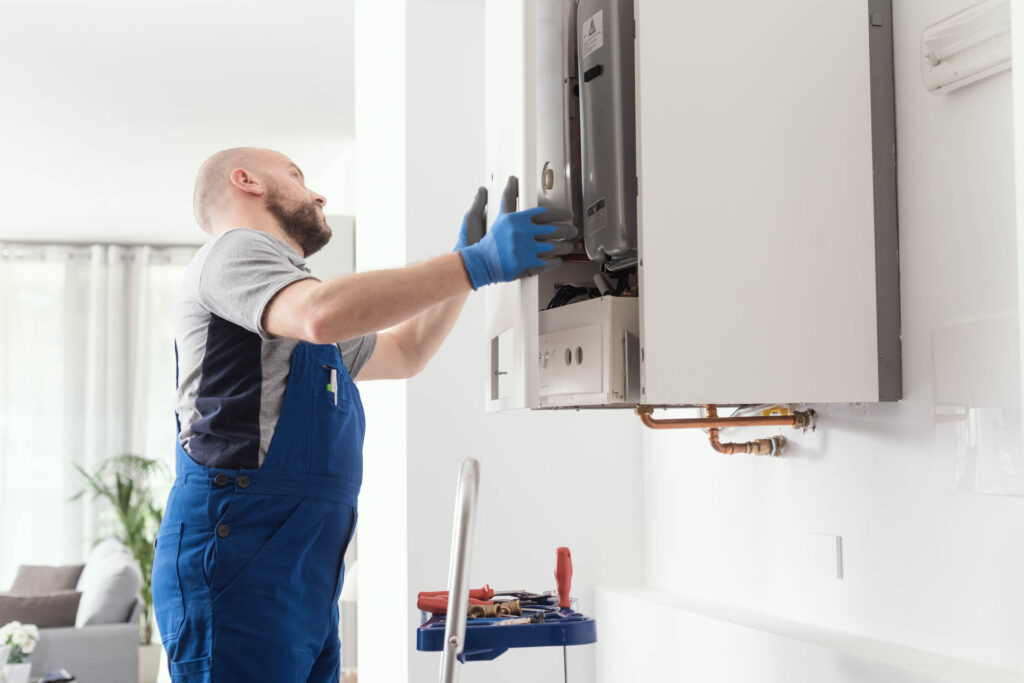 A technician in overalls and gloves services a gas line in a wall-mounted heater in a bright, modern room.