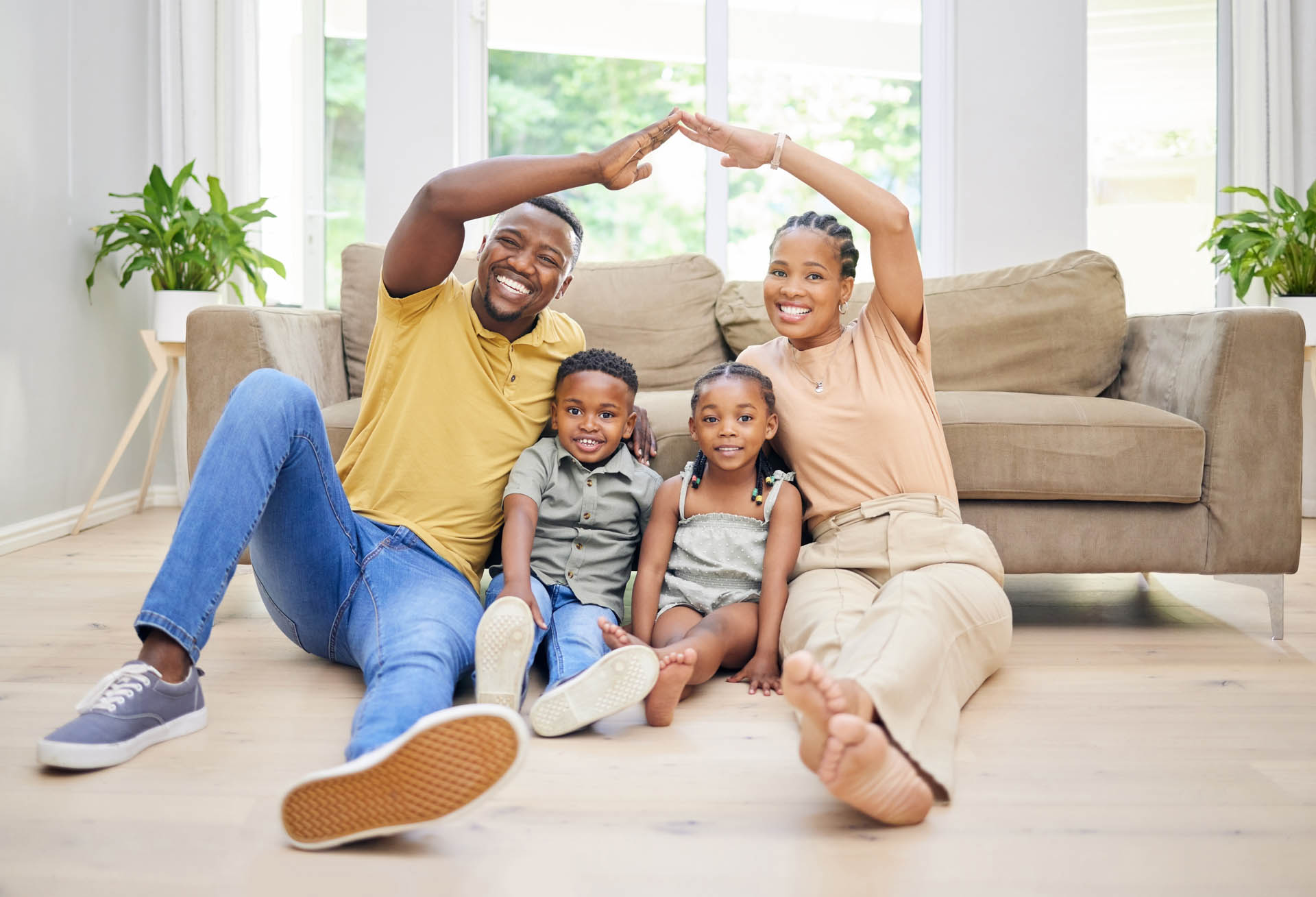 A happy family of four sits on the floor making a roof shape with their arms over two children, smiling in a well-lit living room with visible heating and cooling books on the shelf.