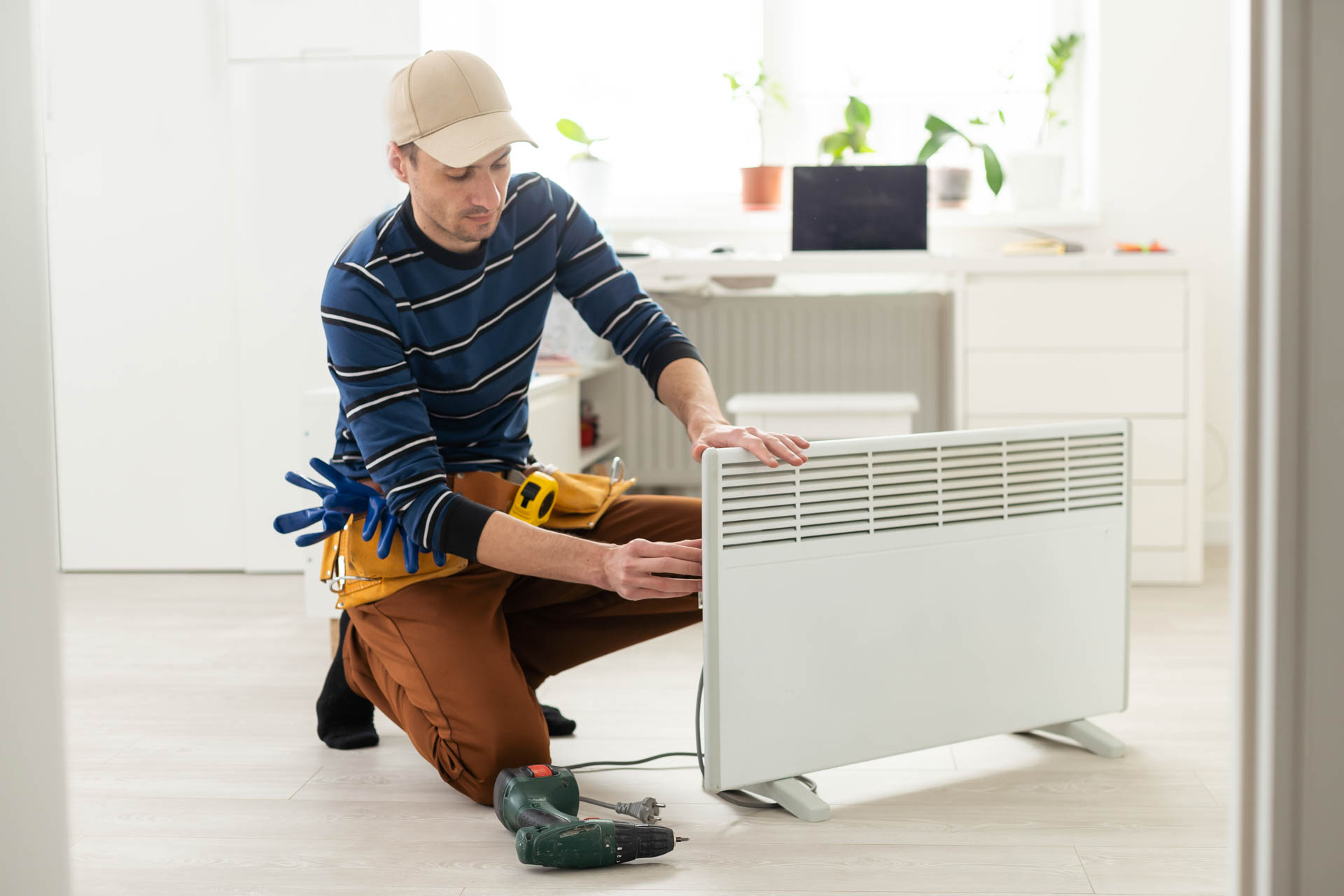 A repairman in a cap and tool belt is kneeling and working on a white portable radiator in a bright room.