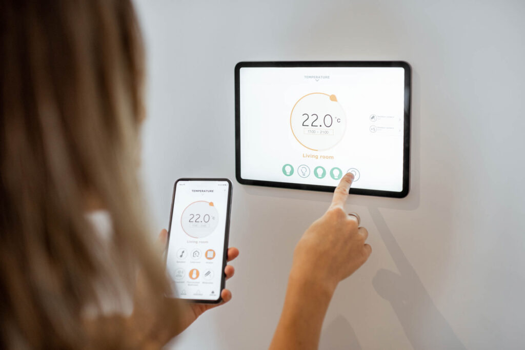 A person adjusts a wall-mounted smart thermostat connected to the gas line using a smartphone app, displaying the same temperature on both devices.