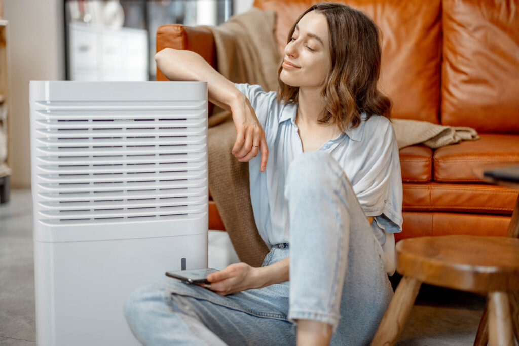 A woman sits on a floor by a dehumidifier, holding a remote and relaxing in a living room with a brown leather sofa near the gas line.