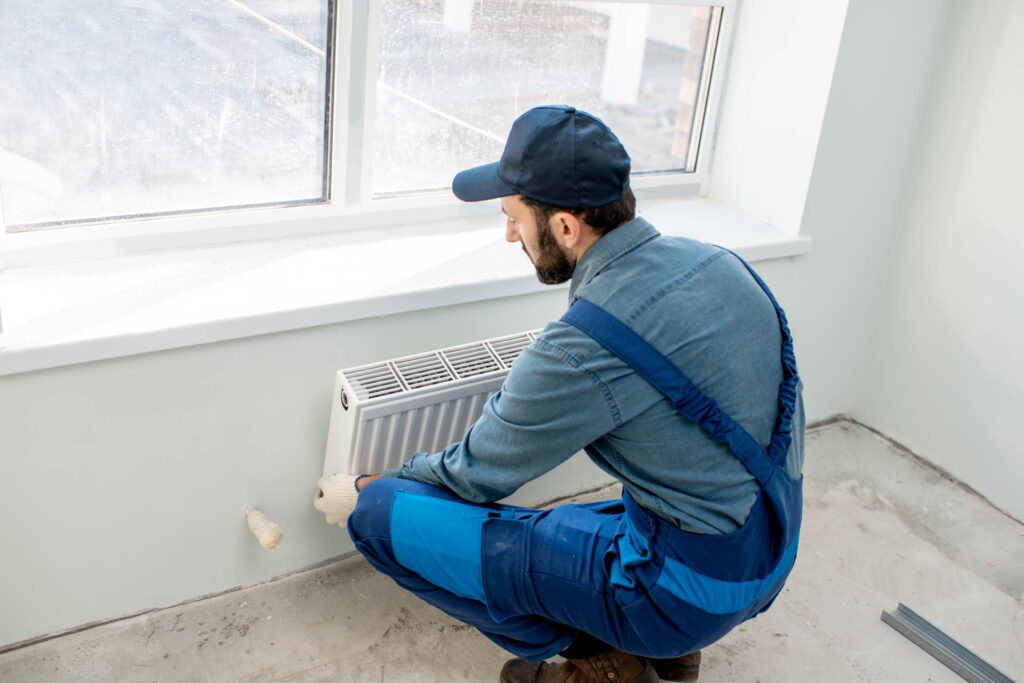 A heating and cooling repairman in blue overalls and a cap is adjusting a radiator in a bright room.