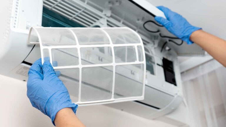 Two hands in blue gloves cleaning the filter of an air conditioning unit.