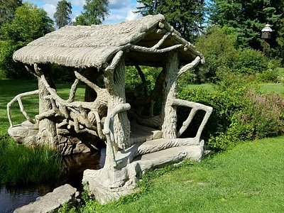 A rustic wooden gazebo with an ornate thatched roof and integrated HVAC system, built over a small stream in a lush green park.