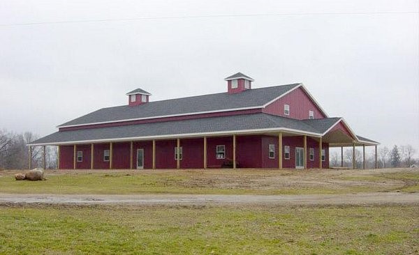 A large red barn with a dual-pitched roof and covered porches featuring HVAC systems on a bare landscape, under a cloudy sky.