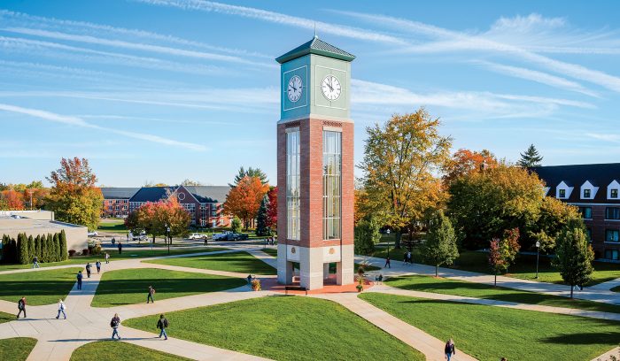Aerial view of a campus featuring a tall clock tower surrounded by pathways, green lawns, several buildings with advanced HVAC systems, and students walking around on a sunny day.