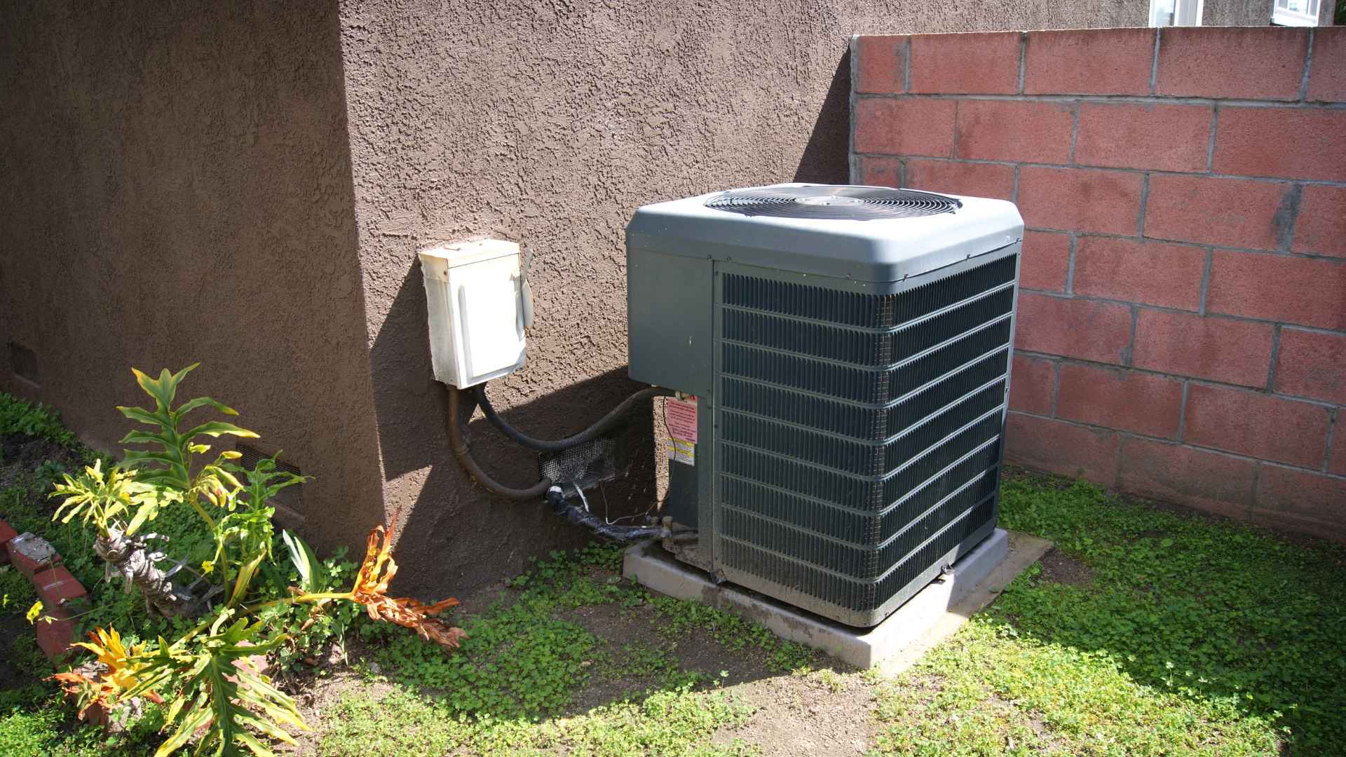 Central air conditioning unit installed on a grassy patch beside a brick wall and a small plant, with exposed electrical wiring and conduit.