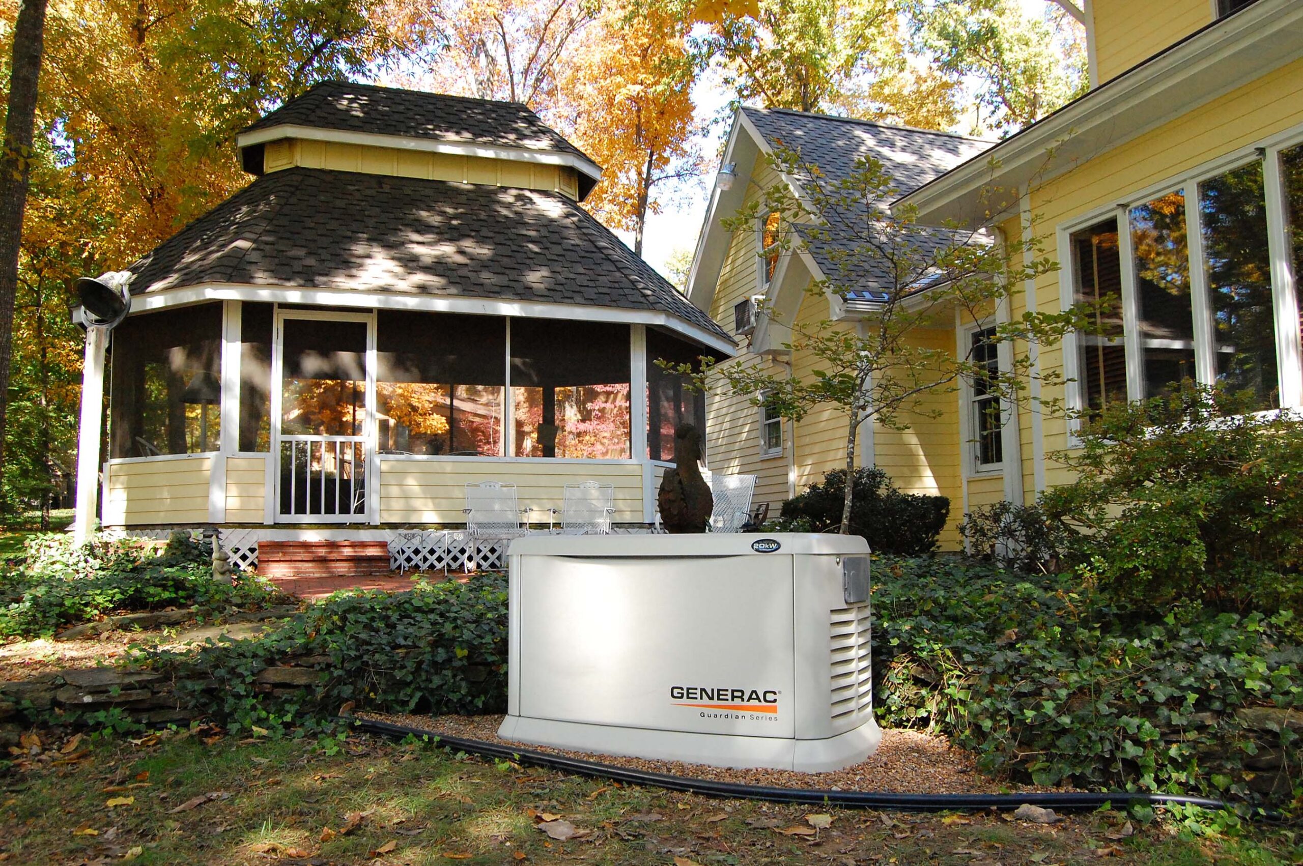A residential backyard featuring a large generac generator near a house with a screened porch surrounded by autumn trees.