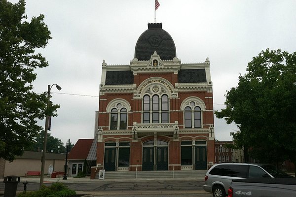 A large brick building with ornate architectural details and a central dome. An American flag is atop the dome. There is a sign near the entrance, and an HVAC unit can be seen near a silver vehicle parked nearby.