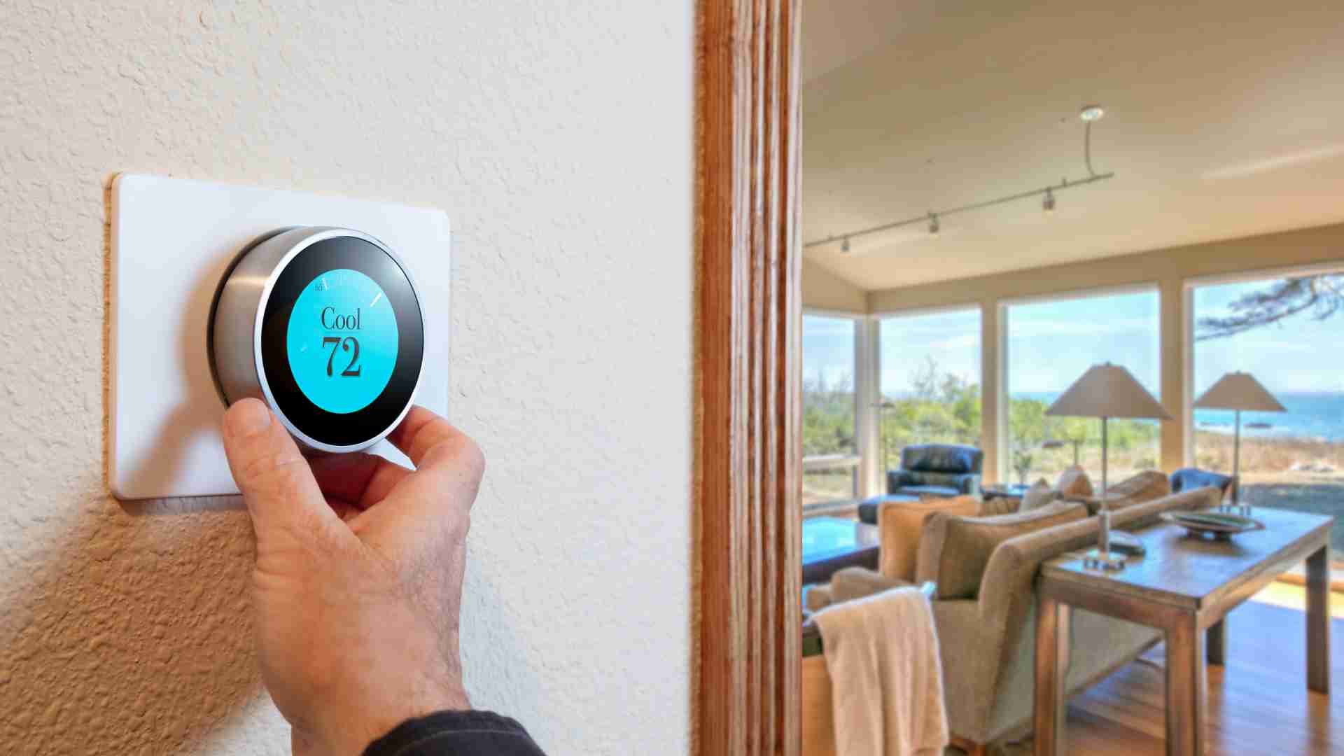 A person adjusts a smart thermostat set to 72°F in a home with a living room view of large windows overlooking a natural landscape.