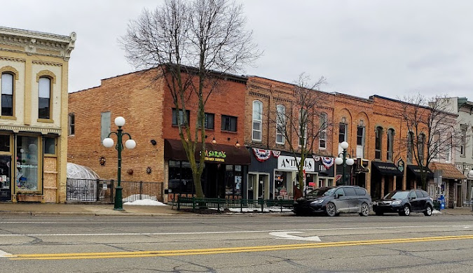 A row of brick buildings with shops and restaurants lines a street with parked cars, lampposts, and leafless trees on a cloudy day.