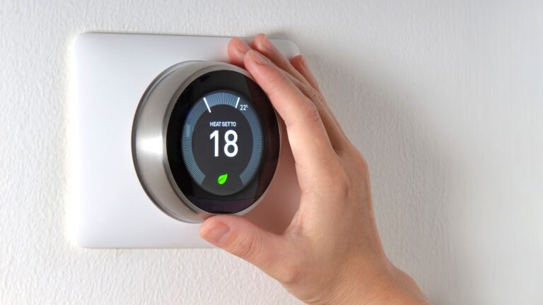 A hand adjusts a smart thermostat on a white wall