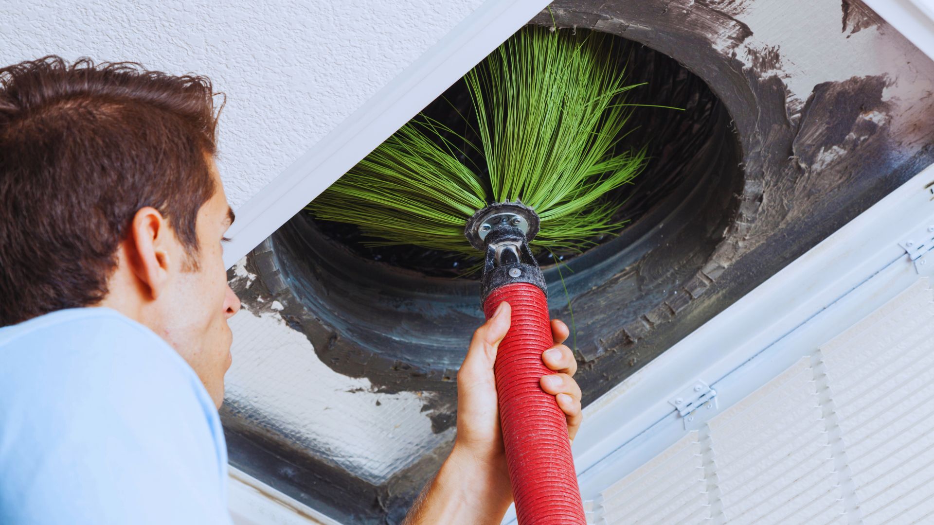 A person uses a rotary brush tool with green bristles to perform duct cleaning inside an air duct in the ceiling.