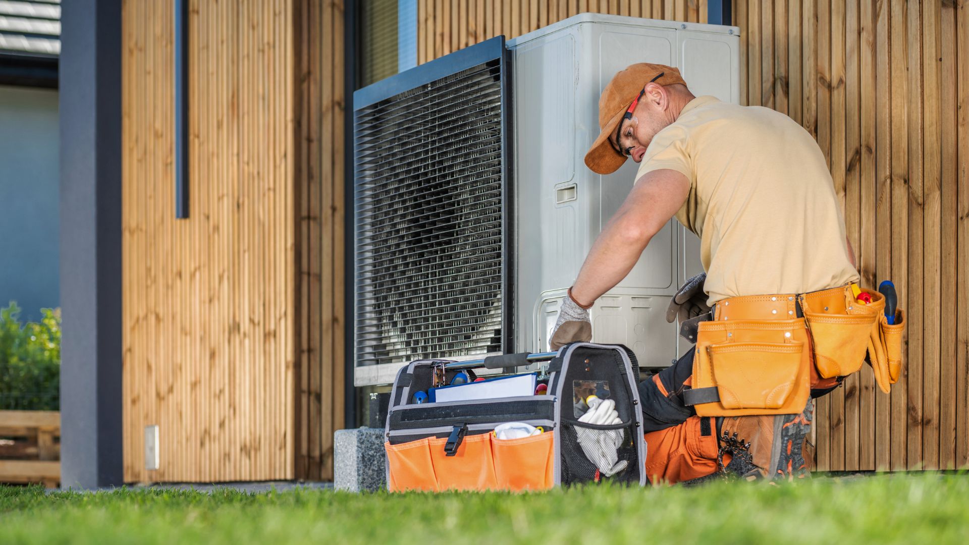heat pump service in michigan A technician wearing an orange hat and tool belt works on an outdoor HVAC unit next to a wooden building, with tools organized in a nearby bag.
