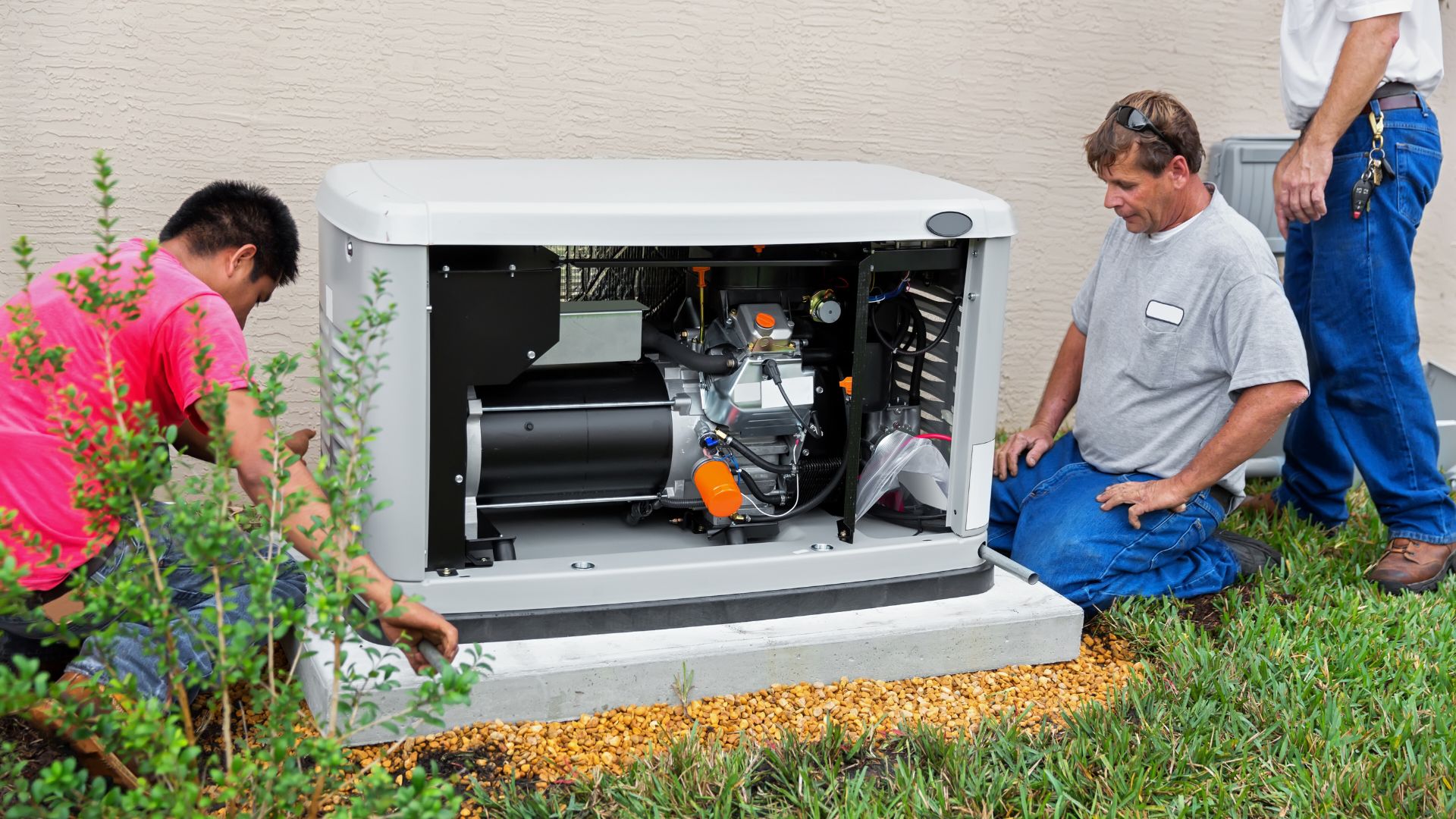home generator installation in michigan Two workers inspect and install a standby generator on a concrete pad next to a beige building, surrounded by grass and small plants.