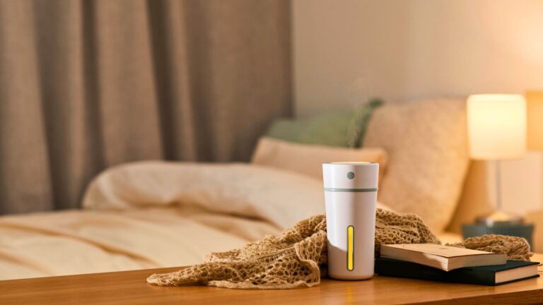 home humidity control in michigan A white humidifier, books, and a knitted blanket sit on a wooden bedside table in a softly lit bedroom with an unmade bed and lamp in the background.