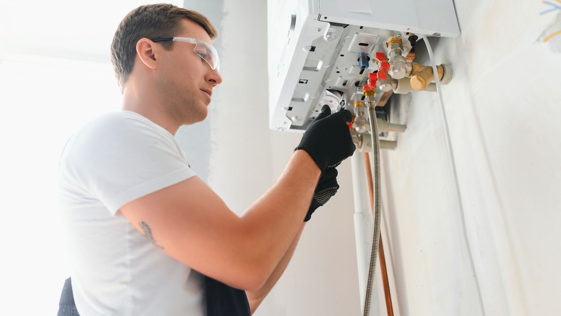 A person wearing safety glasses and gloves adjusts valves on a wall-mounted boiler or water heater.