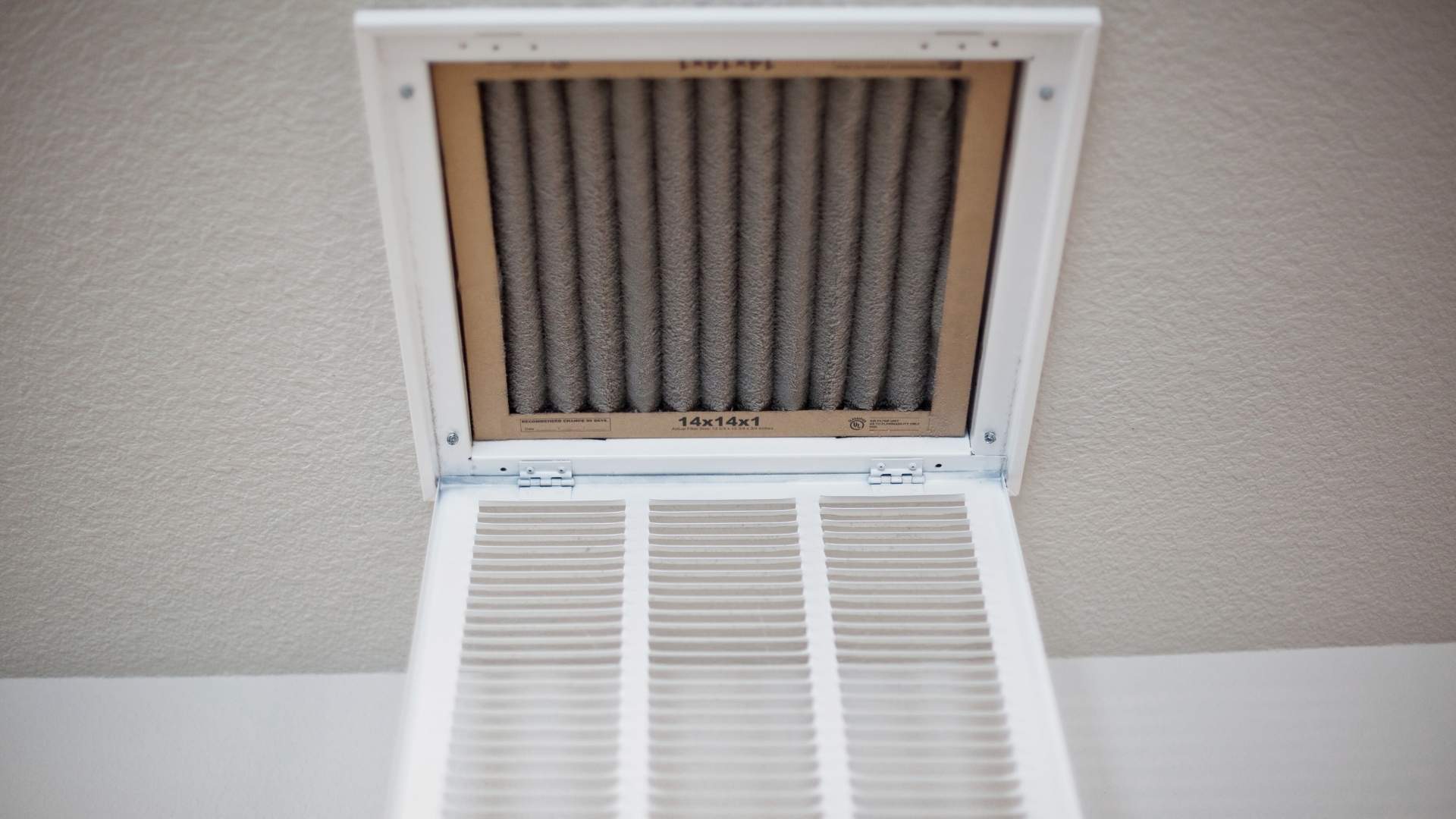 A close-up view of an air vent in a ceiling with its cover open, revealing a dusty air filter inside the vent frame.