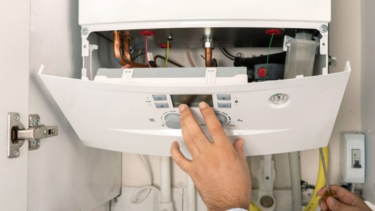 A person adjusts the control panel of an open gas boiler, showing internal pipes and wiring.