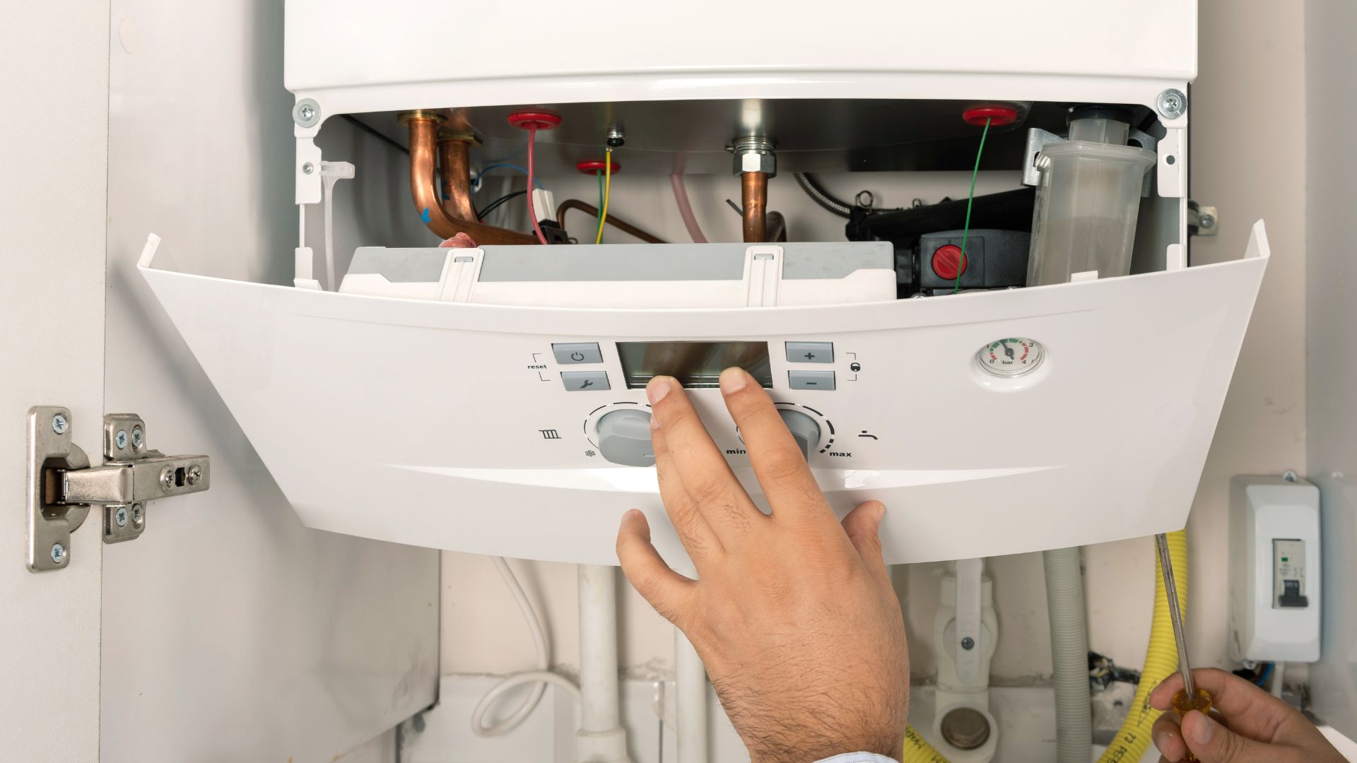 A person adjusts the control panel of an open gas boiler, showing internal pipes and wiring.