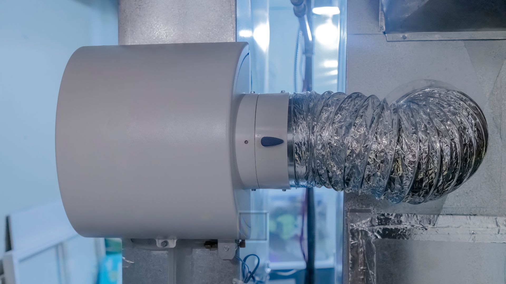 A close-up view of an air duct system with a flexible foil ventilation hose connected to a cylindrical device on a wall.